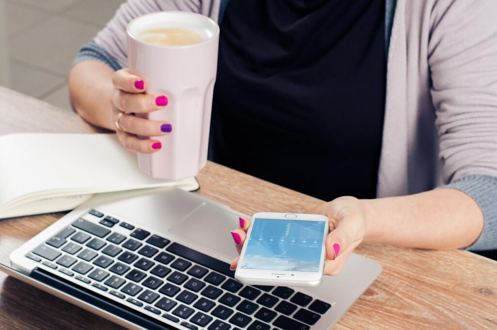 コーヒー片手にパソコンとスマホで仕事をする女性